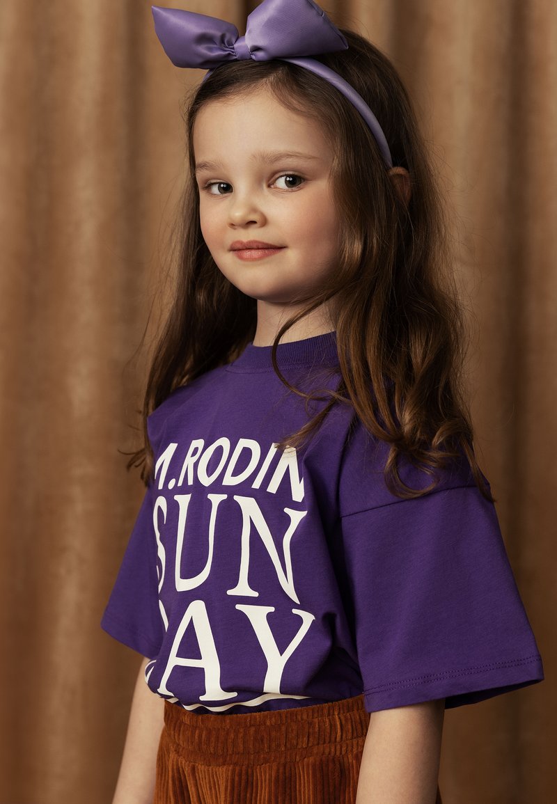 Camiseta de manga corta morada con texto blanco que dice "M. RODIN SUN DAY", combinada con una falda de pana marrón. La niña lleva una diadema morada a juego.