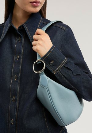 Femme portant une chemise en denim foncé tenant un sac à main en cuir bleu clair avec un détail anneau argenté sur l'épaule sur fond uni.