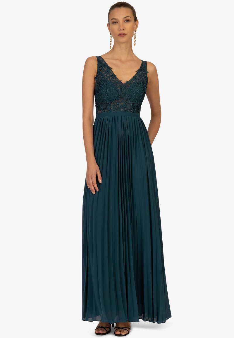 Langes türkisfarbendes Abendkleid mit einem Spitzen-Oberteil und V-Ausschnitt. Der Faltenrock verleiht Textur. Getragen mit schwarzen Riemchenheels, das Model steht frontal.
