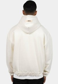 Cream hoodie with a relaxed fit, hood, and elasticized hem. Features a brand label on the back and soft fabric texture.