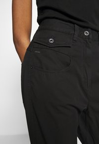 Pantalones cargo negros hechos de tejido resistente, con múltiples bolsillos, cremallera en la bragueta y detalles metálicos. Textura suave con costuras visibles.