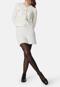 Vit texturerad ullblandning cardigan med guldknappar och ficka, i kombination med en matchande knälång kjol. Svarta ballerinas i kombination med strumpbyxor.