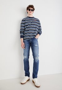 Camisa de rayas azul marino y celeste de manga larga sobre jeans celestes desgastados, combinada con zapatillas blancas con suelas marrones.