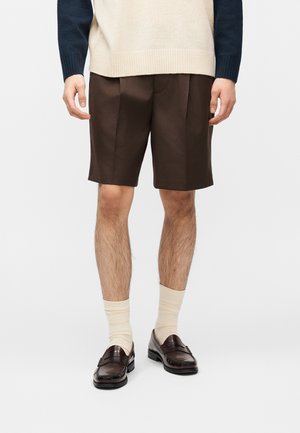 Homme portant un short sur mesure marron foncé longueur genou, des chaussettes crème, des mocassins marron foncé, et un pull beige et bleu marine à manches longues sur fond blanc.