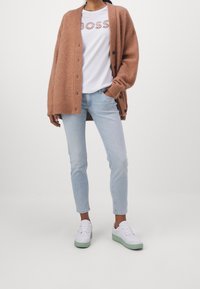 Cárdigan de punto marrón claro sobre una camiseta blanca con impresión "BOSS", combinado con jeans azul claro y zapatillas blancas con suelas verdes.