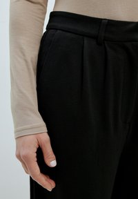 Zwarte, geplooide broek met een gladde textuur, gecombineerd met een lichtbeige lange mouw top. Glad hand rustend naast het kledingstuk.