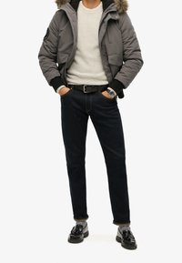 Grijze donsjas met capuchon van namaakbont, witte gebreide trui, donkere spijkerbroek, zwarte riem en glanzende zwarte loafers met kwastjes.