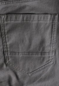 Nærbillede af en tekstureret grå stof baglomme med synlig syning på et beklædningsgenstand, sandsynligvis denimjeans eller lignende materiale.