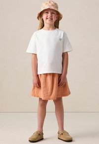 Weißes T-Shirt mit kurzen Ärmeln und grünem Sonnen-Icon, kombiniert mit einem orangefarbenen strukturierten Rock mit floralen Stickereien und beigen Clogs.