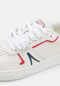 Valge nahast sneaker, millel on kummist põhi, tekstuurilised aktsendid, punased ja sinised detailid ning lamedad valged paelad, mille küljes on perforatsioonid hingavuse tagamiseks.