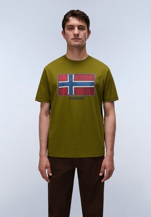 Homme debout face à l’avant portant un t-shirt vert olive avec un motif de drapeau à croix rouge et bleue et un pantalon marron foncé.