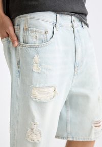 Shorts en denim bleu clair avec des déchirures, présentant des effilochures et une coupe décontractée. Comprend des poches classiques et des passants de ceinture.