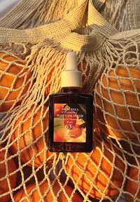 Botella de vidrio ámbar con un tapón gotero blanco, etiquetada como "Suero Aumentador de Vitamina C", colocada en una red color crema con naranjas de fondo.