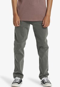 Persona vestida con jeans gris descolorido, zapatillas negras y blancas, y una camiseta malva, de pie con los brazos relajados a los lados.