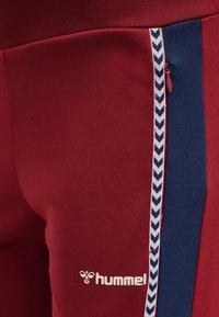 Röda träningsbyxor med en kontrasterande marinblå sidostrand med ett chevronmönster, gummilogo på underbenet och detaljer med dragkedjor.