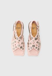 Pink suede sandals with metallic decorative studs, featuring a cross-strap design and open toe. Buckle closure on the side. Square-shaped heel.