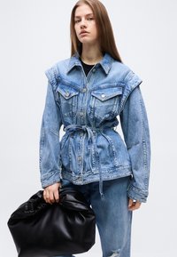 Νεαρή γυναίκα που φοράει τζιν μπουφάν με ζώνη και τζιν, κρατώντας μια μεγάλη μαύρη δερμάτινη τσάντα σε μια απλή λευκή φόντο.