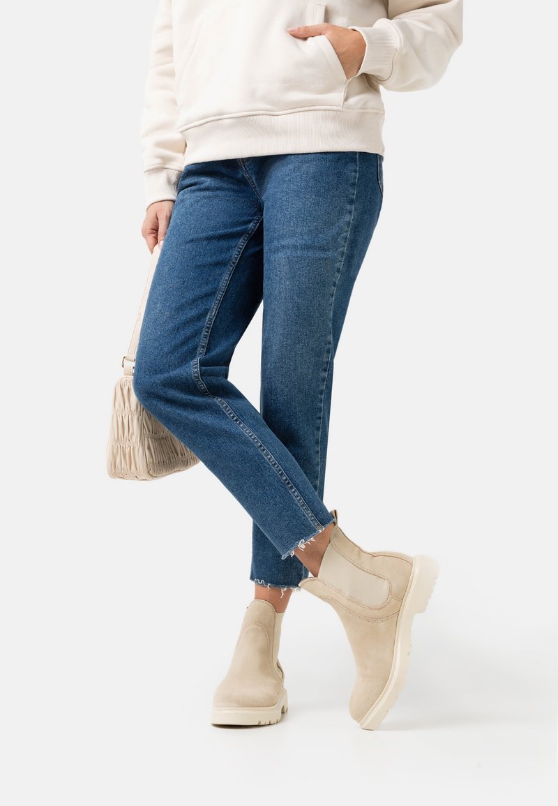 Bottines en suede beige avec des panneaux élastiques sur les côtés, semelles épaisses blanches. Associées à un jean en denim bleu avec un ourlet brut. Petit sac à main tissé.