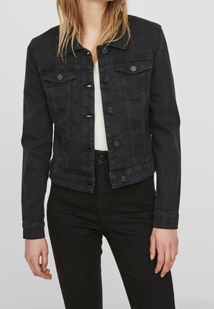 Femme portant une veste en denim noire cintrée avec des fermetures à boutons, des poches poitrine et des manches longues par-dessus un haut blanc et un pantalon noir.