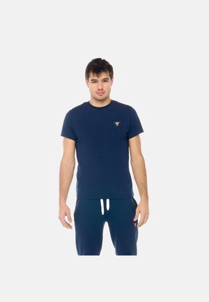 Giovane uomo in piedi rivolto in avanti, indossa una t-shirt blu navy e pantaloni con coulisse abbinati su sfondo bianco.