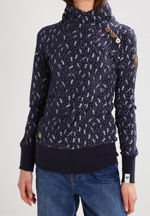 Haut à manches longues bleu marine avec motif hippocampe blanc, trois boutons en bois sur col montant, porté avec un jean bleu.