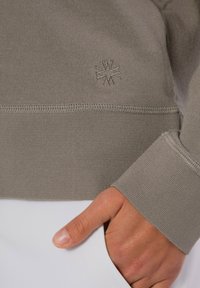 Grauer Sweatshirt mit strukturiertem Design und geripptem Muster, mit einem kleinen geprägten Logo und kontrastierendem Saum. Eine Hand liegt in einer Tasche.