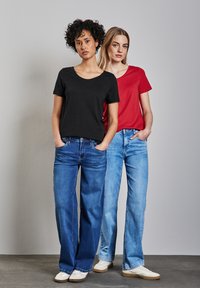 Camisetas de algodón negras y rojas con escote en V, combinadas con jeans de pierna ancha azules. Modelos usando zapatillas blancas sobre un fondo neutro.