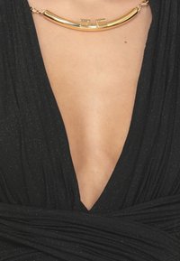 Indumento nero e texturizzato con scollatura a V profonda, adornato con un collier dorato lucido. Il tessuto ha una leggera luminescenza.