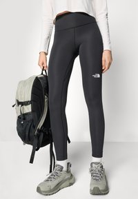Sorte leggings med høj talje, prydet med et lille hvidt logo. Kombineret med en lysegrå rygsæk og grå vandresko.