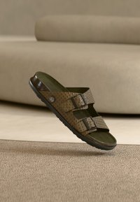 Sandale marron texturée à double bride avec boucles noires flottant au-dessus d'un tapis beige, avec un canapé beige assorti en arrière-plan.