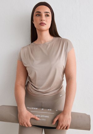 Jeune femme portant une tenue de sport beige et un bandeau, tenant un tapis de yoga beige roulé contre un mur blanc uni.