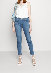 Camiseta de satén blanca con encaje, combinada con jeans de color azul claro. Sandalias de plataforma blancas con tiras entrecruzadas. Bolso de hombro azul con patrón.