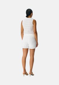 Top sin mangas de encaje blanco y pantalones cortos a juego, con un patrón texturizado. Combinado con mules de tacón en un color claro.