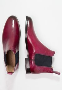 Pár lesklých burgundských kožených Chelsea bot s černými elastickými bočními panely a poutky na bílém pozadí.