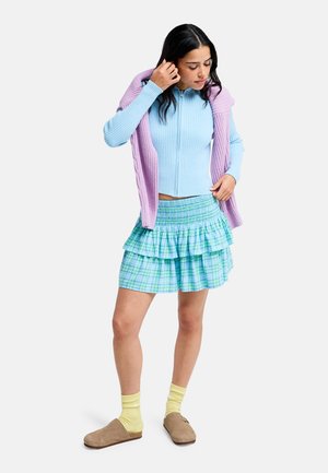 Jonge vrouw die een lichtblauwe trui met rits draagt, een paarse trui als laag over de schouders gedrapeerd, een blauw geruite rok, gele sokken en bruine instapschoenen.
