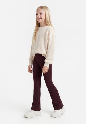 Smilende pige med langt blondt hår iført en cremefarvet strikket sweater, mørkelilla bukser og hvide sneakers, stående mod en hvid baggrund.