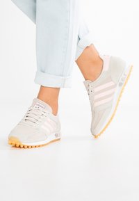 Světlé béžové sportovní boty s růžovými akcenty, materiály síťoviny a semiše, kulatá špička, strukturovaná podrážka a tři bílé proužky na straně.