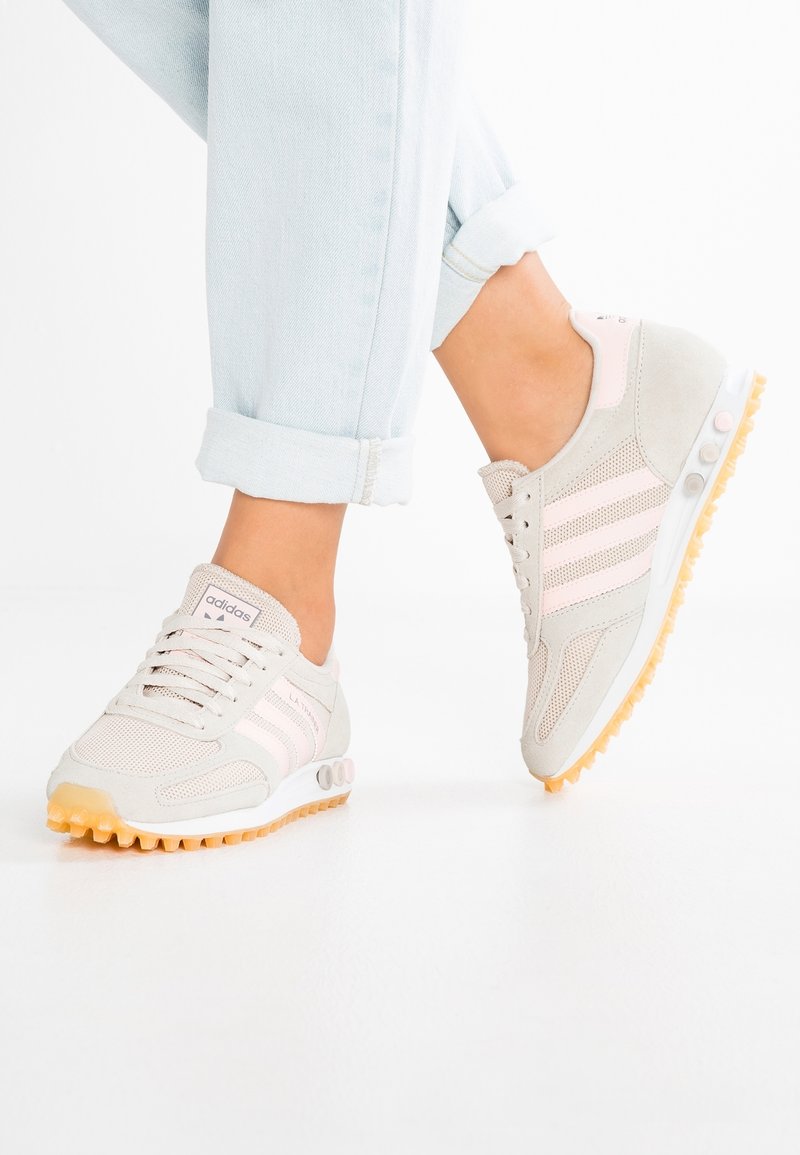 Světlé béžové sportovní boty s růžovými akcenty, materiály síťoviny a semiše, kulatá špička, strukturovaná podrážka a tři bílé proužky na straně.