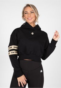 Sudadera corta negra con capucha ajustable, con detalles en beige y una marca en las mangas. Tejido suave, corte relajado.