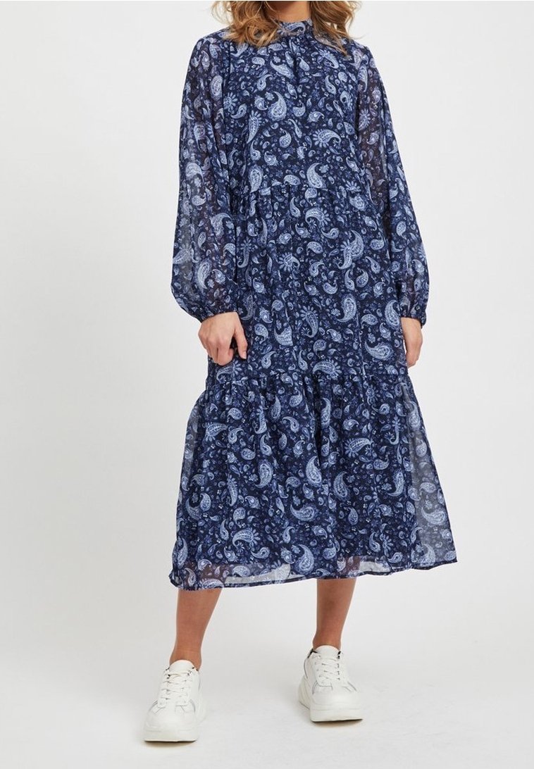 Femme portant une robe à manches longues bleu marine à motif cachemire avec une jupe à volants et des baskets blanches, debout devant un fond uni.