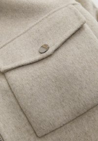Gros plan sur une poche de manteau en laine beige avec rabat fermé par un bouton-pression rond argenté.