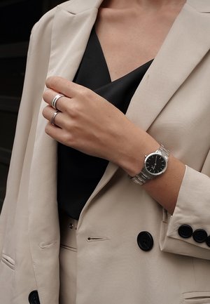 Femme en costume beige et top noir portant une montre argentée au poignet gauche et deux bagues argentées aux doigts de la main droite.