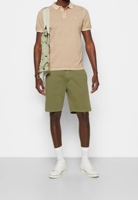 Beige pikétröja med korta ärmar och broderad logotyp, matchat med gröna shorts, vita sneakers och en ljusgrön axelväska.