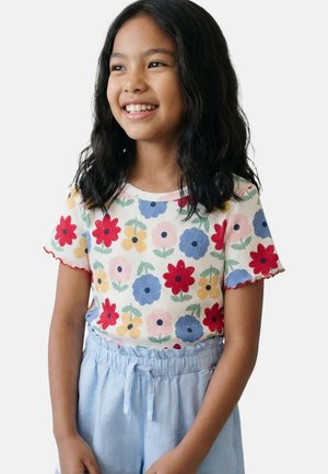 Fille souriante aux longs cheveux foncés portant un haut blanc manches courtes avec des fleurs colorées et un pantalon taille haute bleu clair avec ceinture à nouer.