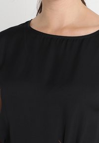 Mujer que lleva una blusa negra sencilla con escote redondo, que muestra su cuello y la zona de la clavícula.