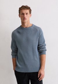 Lichtblauwe gebreide trui met een textuurpatroon, ronde hals en geribde manchetten. Het model draagt donkere broeken, afgezet tegen een neutrale achtergrond.