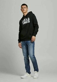 Black hooded sweatshirt with white lettering, blue distressed jeans, and white sneakers. Model stands against a plain background.