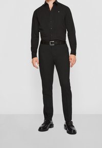 Chemise noire à manches longues avec col boutonné, associée à un pantalon noir sur mesure. Comprend un logo discret et des chaussures en cuir noir.
