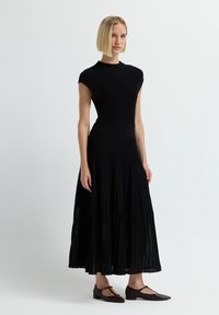 Abito midi nero plissettato con corpino aderente e maniche a cappuccio, realizzato in tessuto testurizzato, abbinato a scarpe basse marrone scuro.
