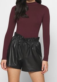 Viininpunainen pitkähihaisen pusero, jossa on mock-kaulus, yhdistettynä mustiin tekonahkashortseihin, joissa on rypytetty joustava vyötärönauha ja nyöri.